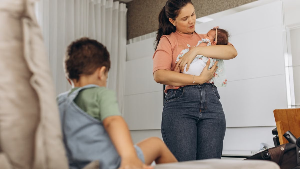 A mother lovingly cradles her baby while her child sits nearby on the couch in a modern room setting.