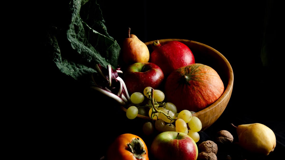 Dark still life composition featuring assorted autumn fruits in a wooden bowl, creating a moody and artistic scene.